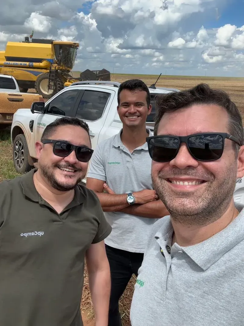 Tres integrantes de upCampo sonríen en el campo, con una cosechadora al fondo
