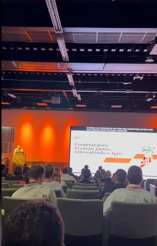 Orador en el escenario con la pantalla que proyecta "Cooperar para avançar juntos, reinventando o Agro" (Cooperar para avanzar juntos, reinventando el agro)
